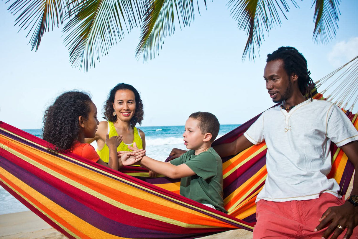 Family of four in a colourful hammock on a beach with palm trees.