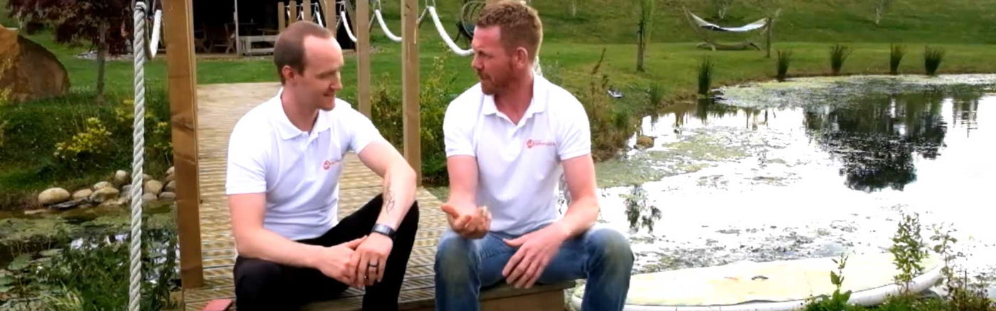 Two men in white shirts sitting on a swing by a pond