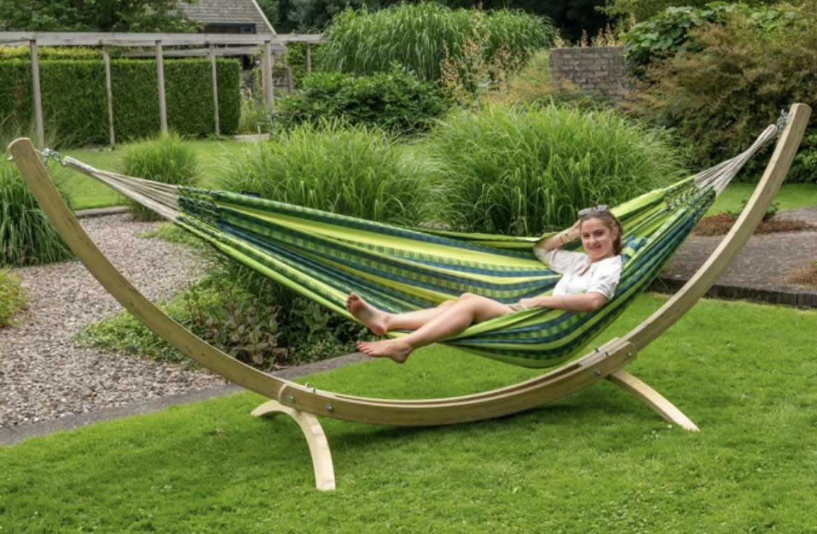 Person relaxing in a green and blue striped hammock with a wooden stand on grass.