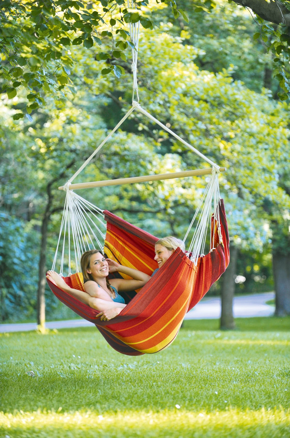 Two people are relaxing in a colourful hammock hanging between trees in a park.