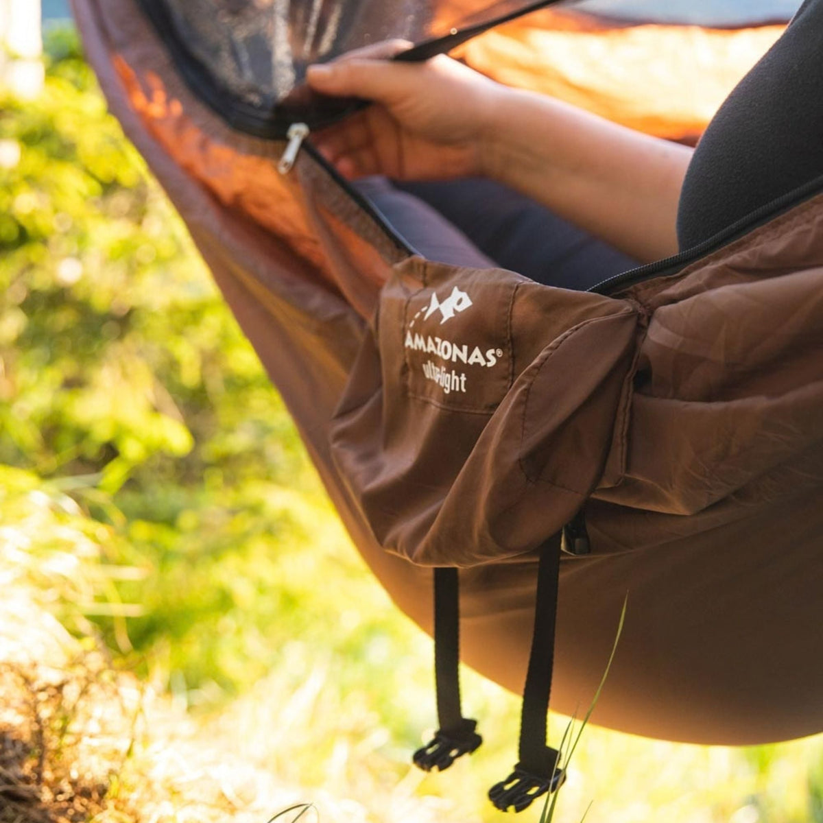 A person's hand and leg are shown inside a brown hammock outdoors, with the brand name 'AMAZONAS ultra-light' and logo visible on the side pocket.