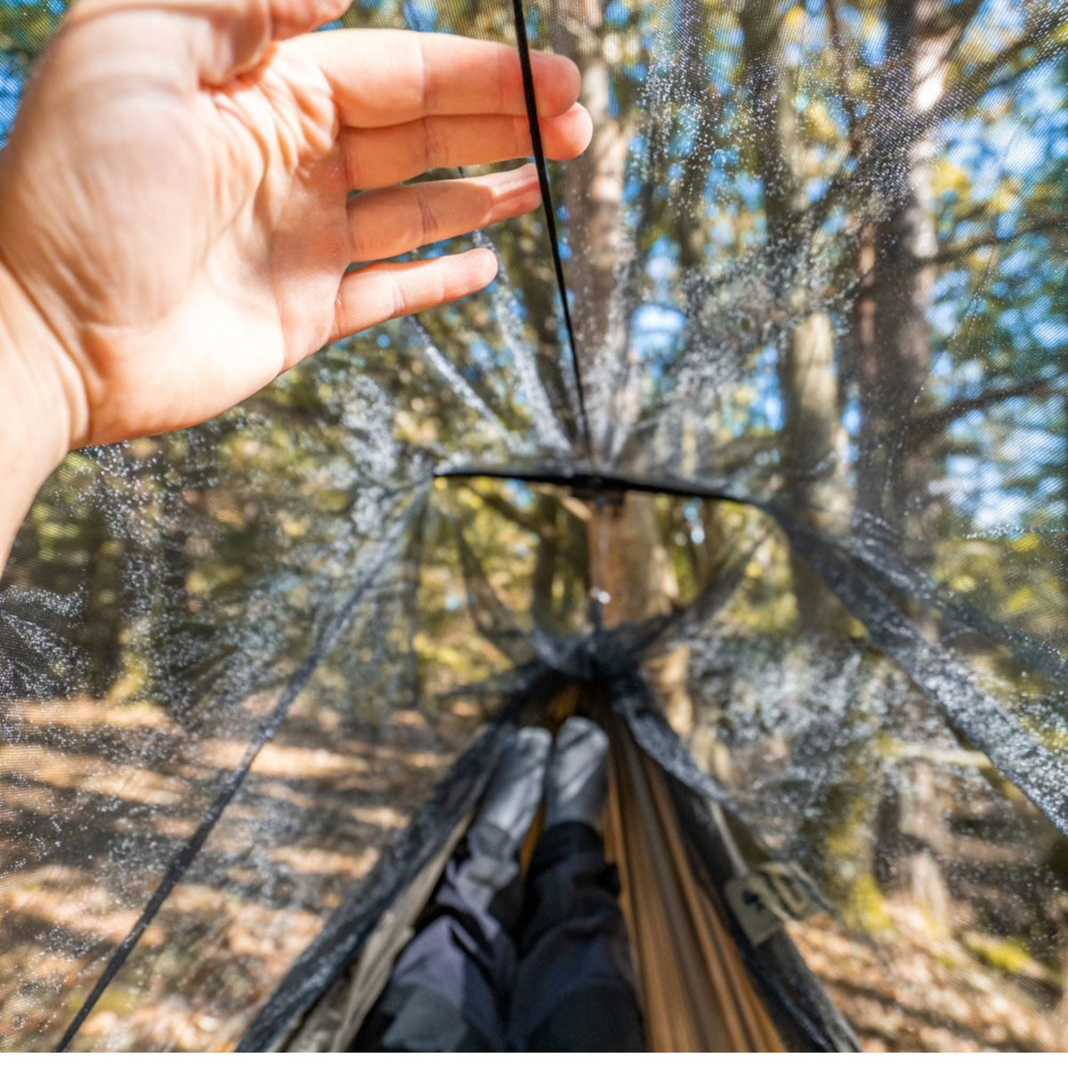 A transparent rainfly net is shown in a forest setting, with a person's hand touching the mesh material and their legs visible inside the hammock below.