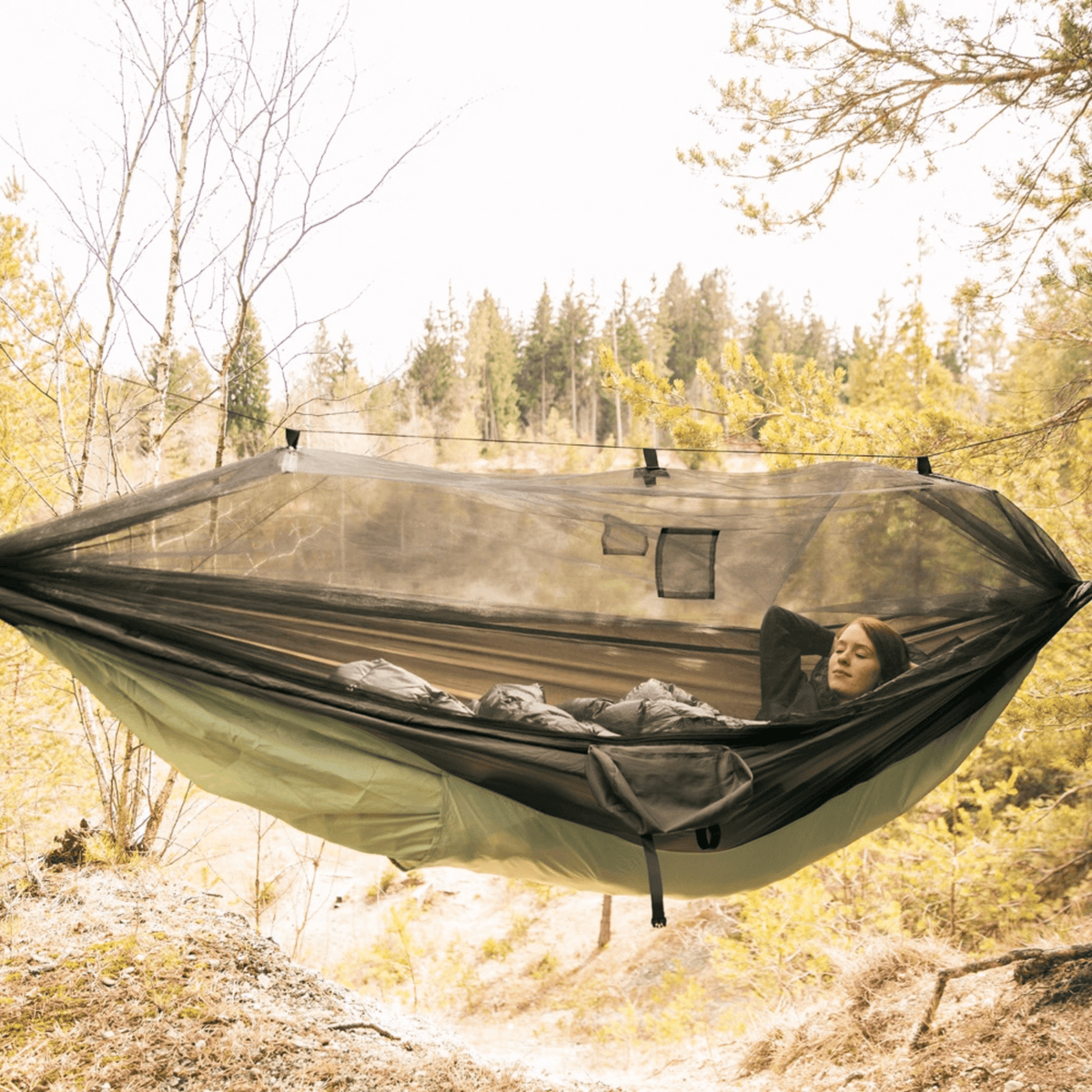 A person relaxes in a green and black mesh camping hammock suspended between trees in a forest setting.
