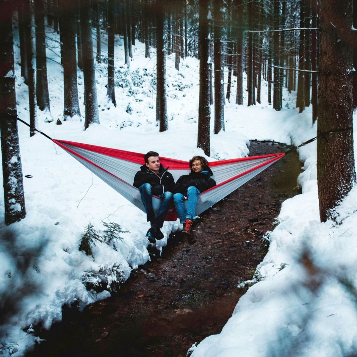 A red and white camping hammock suspended between trees in a snowy forest, with two people sitting inside relaxing.