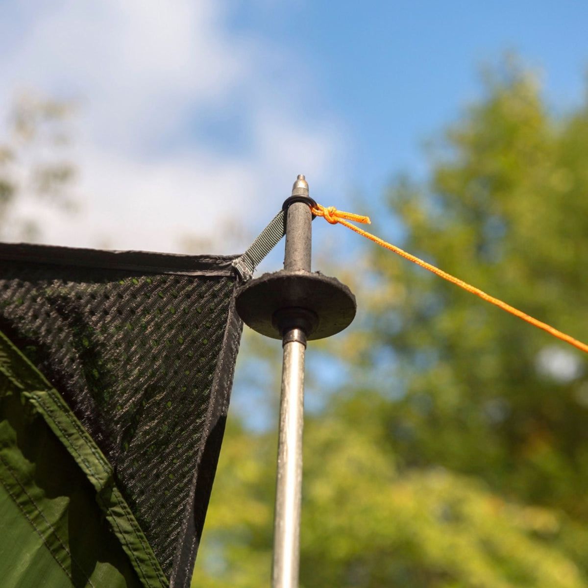 Close-up of a tent pole with a black circular base and an orange guy line attached to a mesh fabric, photographed outdoors against a blurred blue sky and green trees.