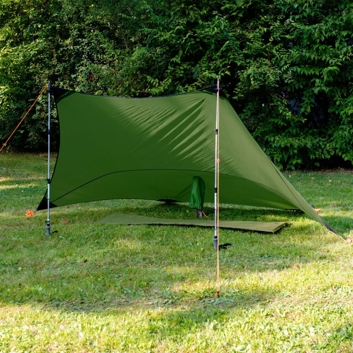 An olive green tarp shelter set up on a grassy lawn with trees in the background, supported by two poles.