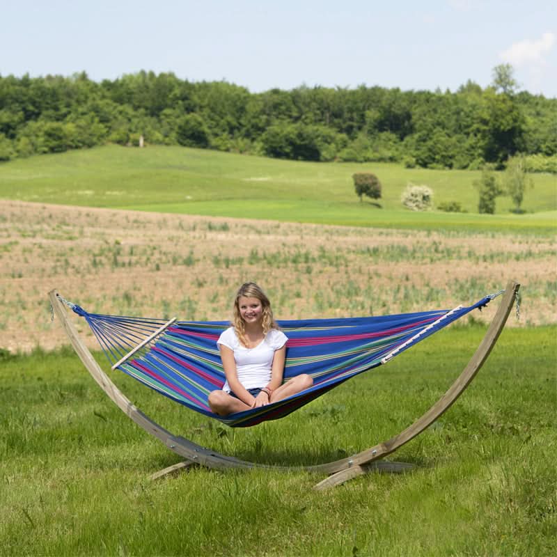 A woman sitting in a colourful hammock with a wooden stand in a grassy field.