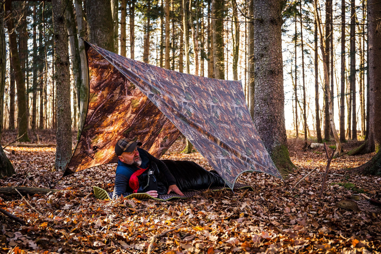 Person lying under a camouflage tarp in a forest setting