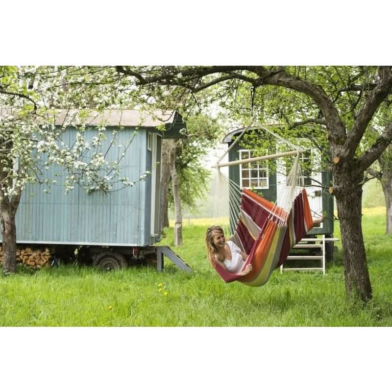 Person relaxing in a colourful hammock under a tree with a small wooden cabin in the background.