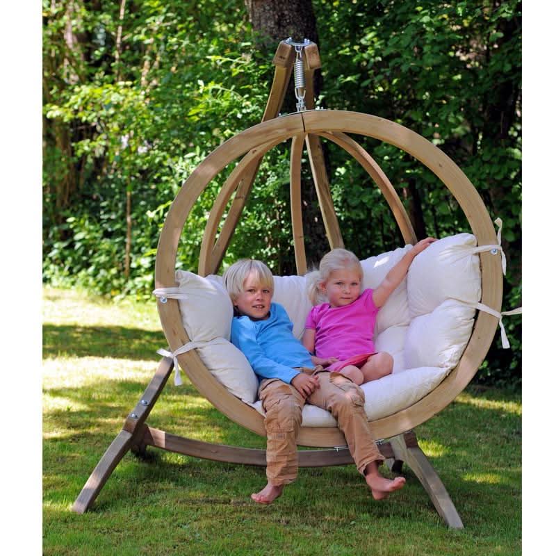 Two children are sitting in a wooden hanging chair outdoors.