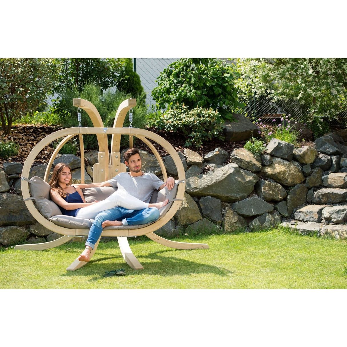 Two people are sitting in a circular wooden swing chair in a garden setting.