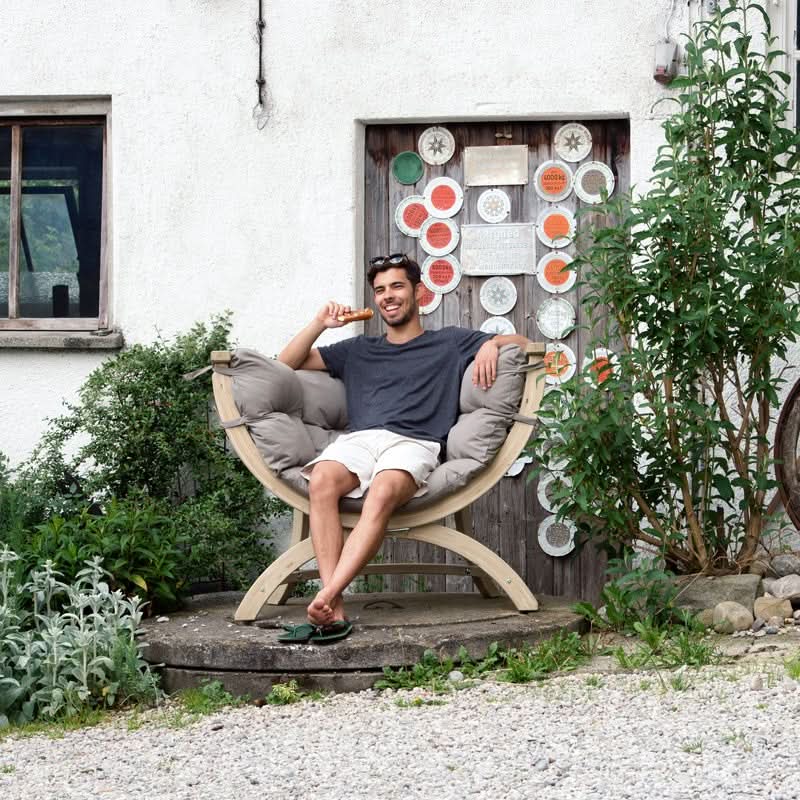 Man sitting in a decorative chair outdoors with a white building and plants in the background