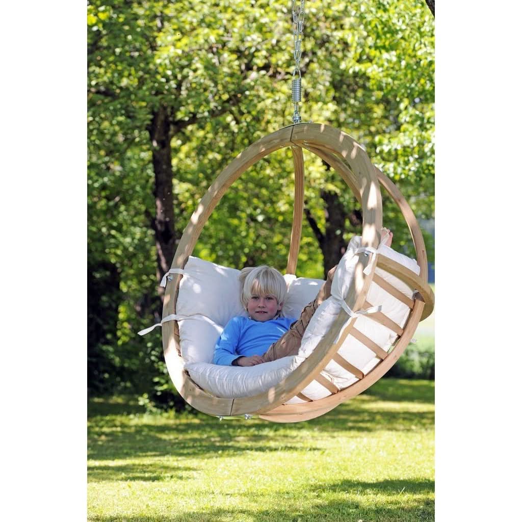 Child in a hanging chair with a white cushion and a wooden frame, surrounded by greenery.