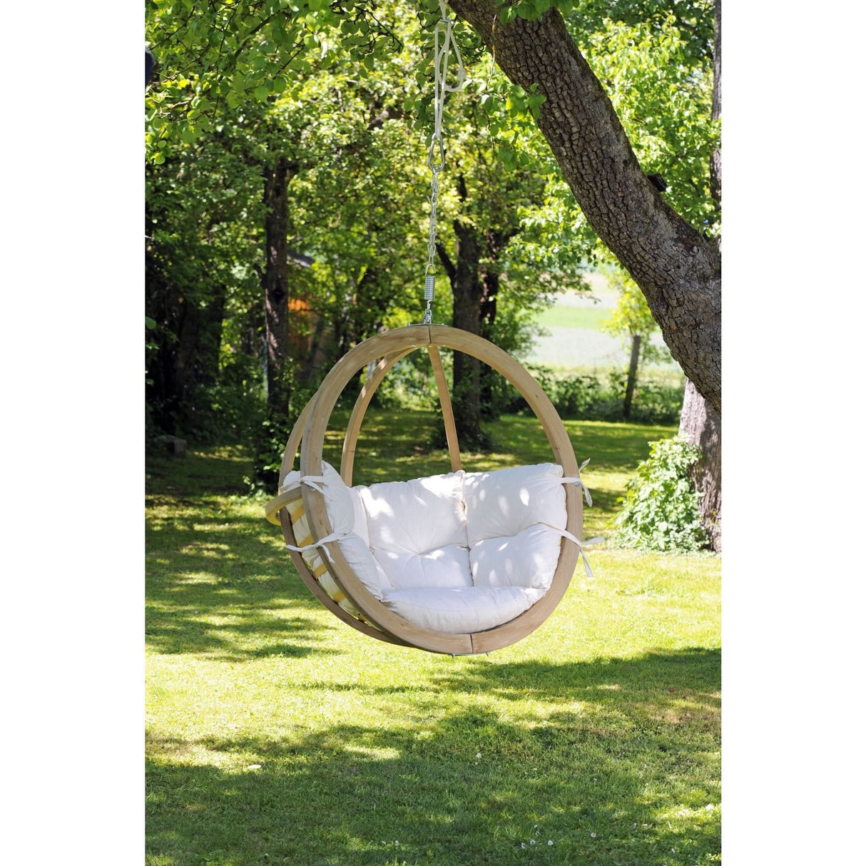 A hanging chair with white cushions hanging from a tree in a garden.
