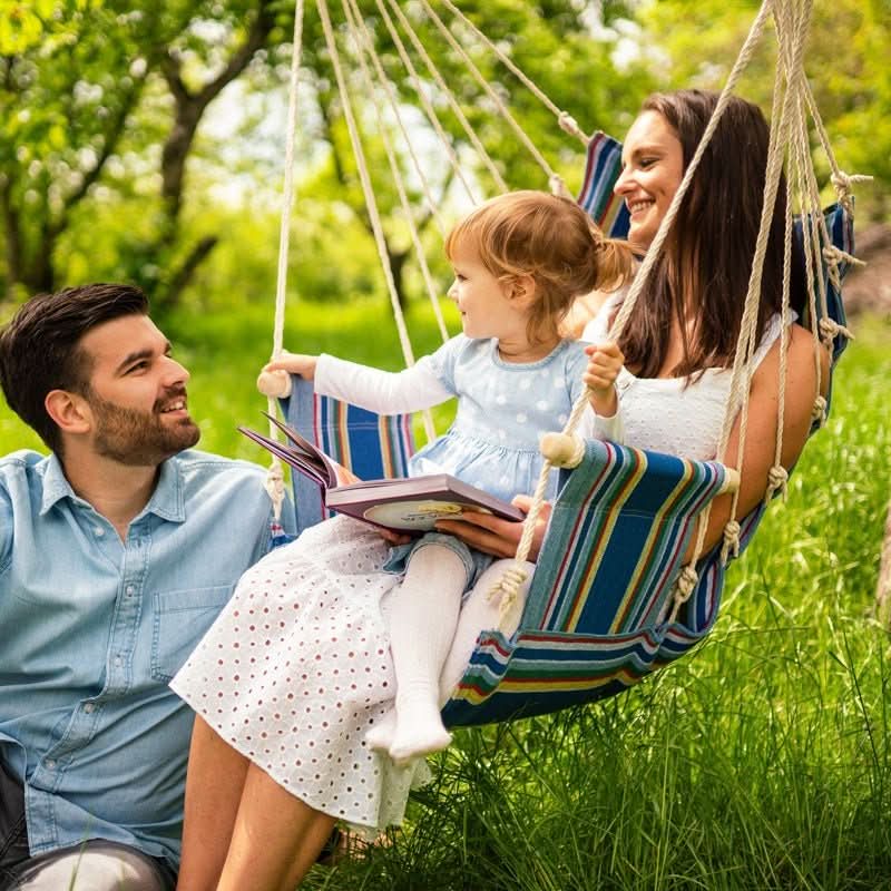 Family of three in a striped hammock with a book, surrounded by greenery