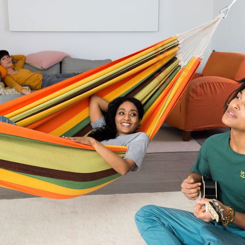 A woman in a colourful striped hammock with a person playing guitar nearby in a living room setting.