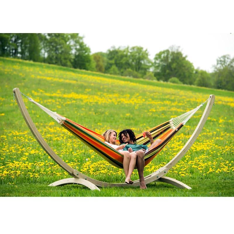 Two people are relaxing in a colourful hammock with a wooden stand in a field of yellow flowers.