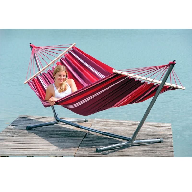 Person relaxing in a red hammock with a stand on a wooden deck by a body of water.
