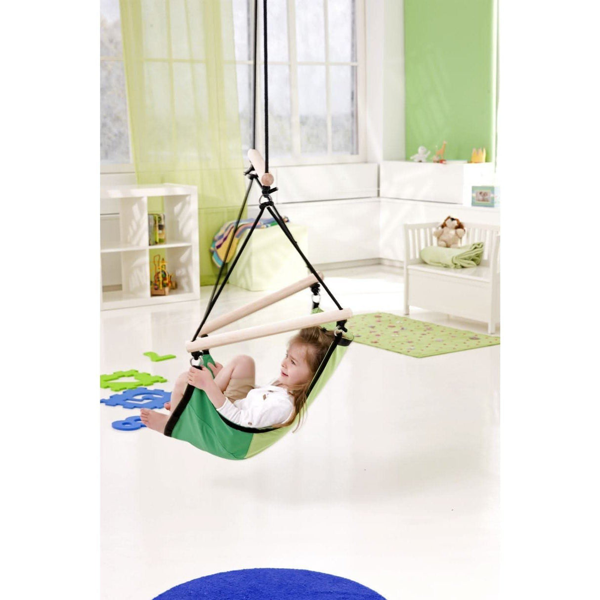 Child in a green indoor swing in a bright room with white walls and green accents.