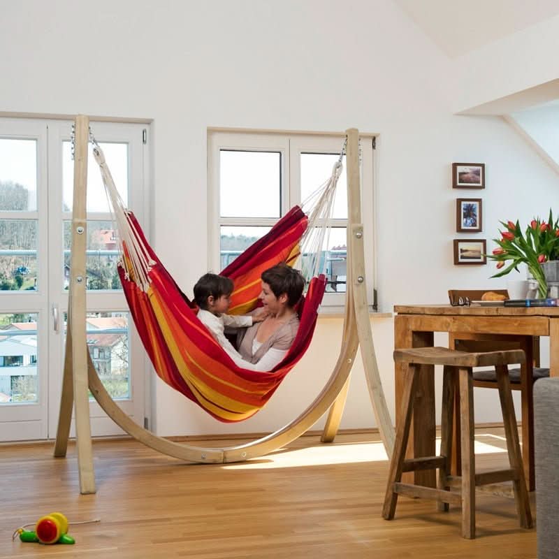 Two people in a colourful striped hammock inside a room with wooden furniture and large windows.