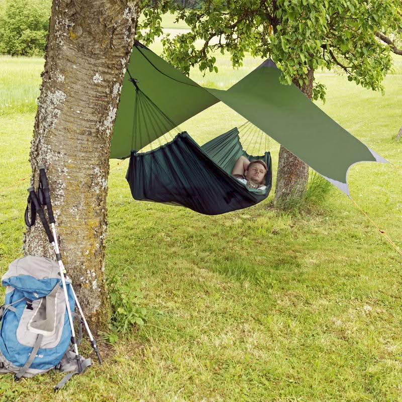 Person relaxing in a green hammock between two trees with a backpack nearby.
