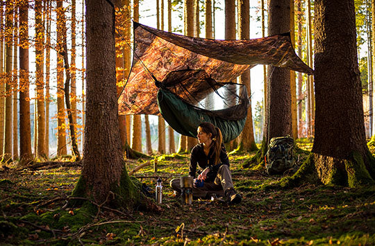 A woman sat in a forest with a green hammock underquilt and a camouflage hamock tarp.