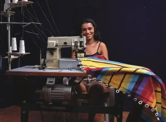 Woman sitting at a sewing machine with a colorful fabric draped over it against a dark background