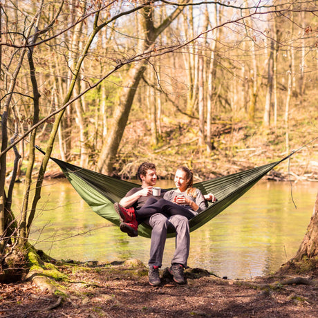 Two people sitting in a green hammock by a river in a forest.