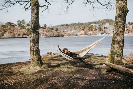 Person relaxing in a hammock by a lake with trees and houses in the background