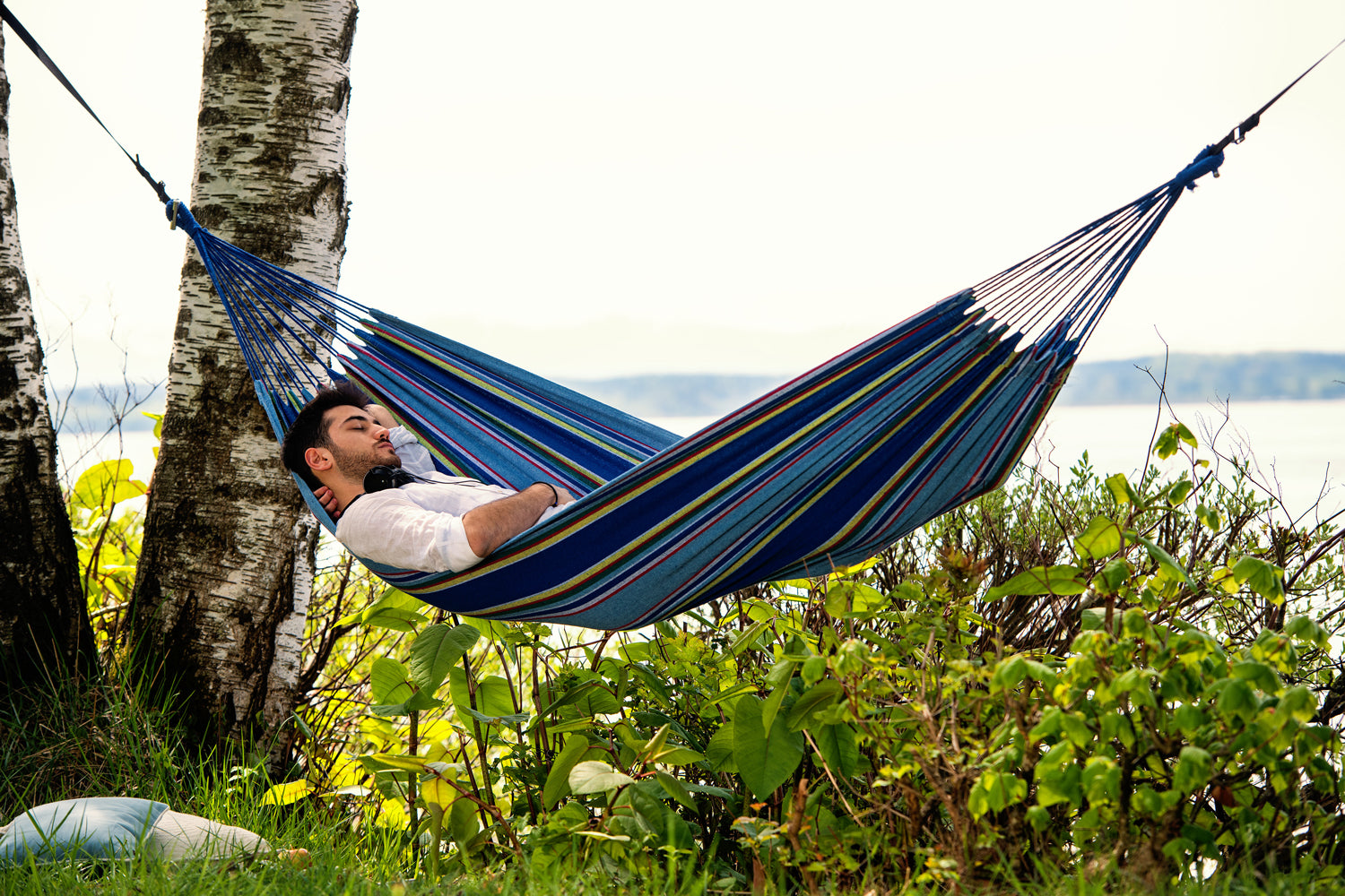 Man relaxing in a blue striped hammock by a lake with trees and grass in the background