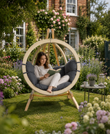 Woman reading a book in a circular wooden chair surrounded by flowers and greenery.