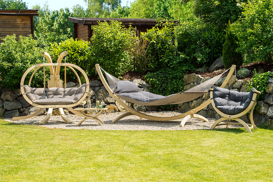 A serene garden scene with a hammock and cushioned chairs on a gravel area, surrounded by lush greenery and a stone wall, radiates relaxation.