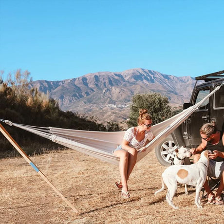 Woman sitting in a hammock with a dog and another person near a vehicle with mountains in the background.