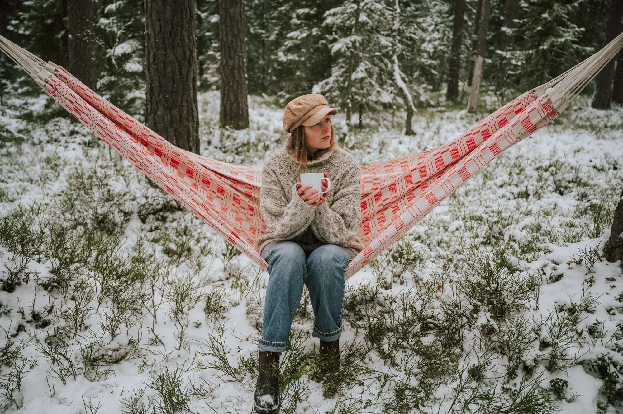A woman in a cosy sweater and cap sits on a red checkered hammock in a snowy forest, holding a mug. The scene conveys tranquillity and warmth.