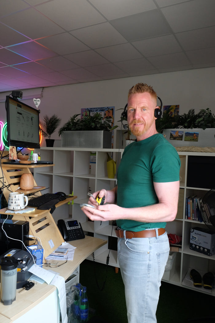 A man in a green shirt is standing in an office with shelves and computer equipment.