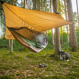 A person relaxes in a green camping hammock suspended beneath a large tan tarp shelter in a forest. A backpack, cup, and shoes sit on the mossy forest floor nearby.