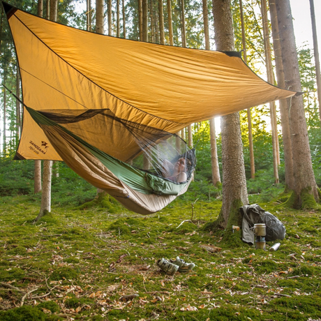 A person relaxes in a green camping hammock suspended beneath a large tan tarp shelter in a forest. A backpack, cup, and shoes sit on the mossy forest floor nearby.