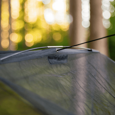 Close-up of a dark gray mesh canopy or hammock with a black hanging cord and a flexible spreader bar, photographed outdoors with a blurred forest background.