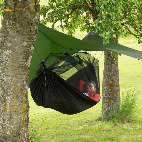 A black mosquito net hammock suspended between two trees, with a person lying inside under a green tarp canopy in an outdoor grassy setting.