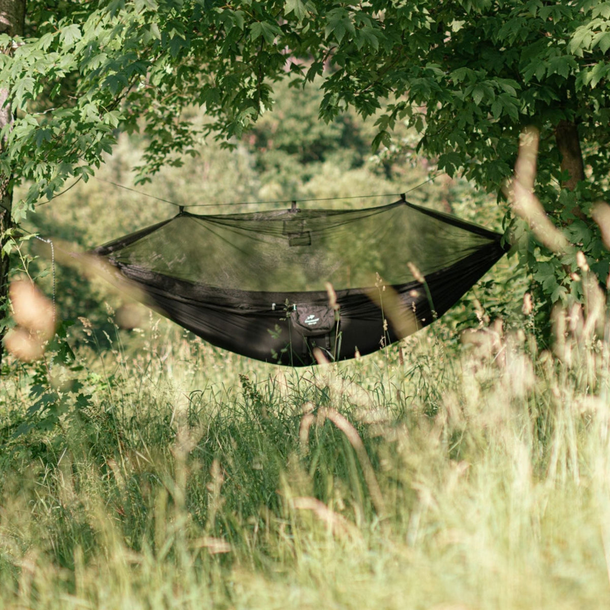 A black camping hammock with an attached mosquito net suspended between two trees in a grassy, wooded outdoor setting.