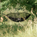 A black camping hammock with an attached mosquito net suspended between two trees in a grassy, wooded outdoor setting.