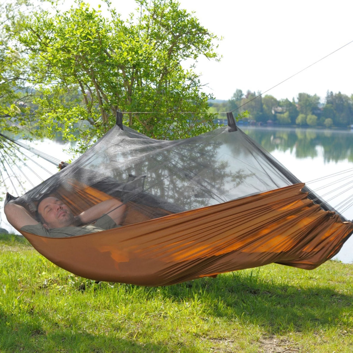 A person relaxes in a brown hammock suspended outdoors over a lake, with a black mosquito net draped over the hammock and trees and water in the background.