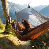 A woman relaxes in a brown hammock suspended between two trees, with a black mesh canopy overhead and a scenic mountain landscape in the background.