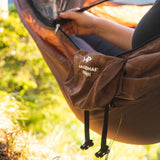 A person's hand and leg are shown inside a brown hammock outdoors, with the brand name 'AMAZONAS ultra-light' and logo visible on the side pocket.
