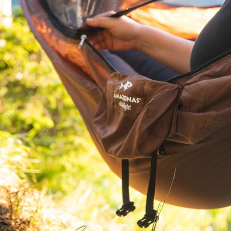 A person's hand and leg are shown inside a brown hammock outdoors, with the brand name 'AMAZONAS ultra-light' and logo visible on the side pocket.