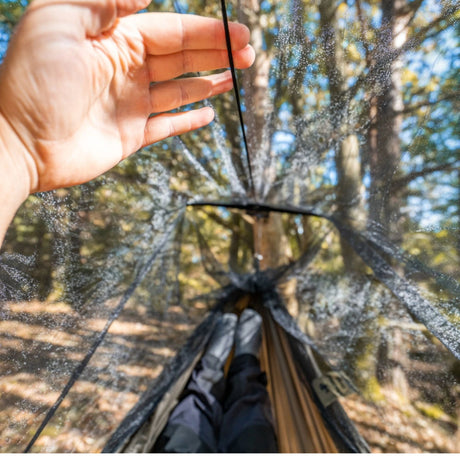 A transparent rainfly net is shown in a forest setting, with a person's hand touching the mesh material and their legs visible inside the hammock below.