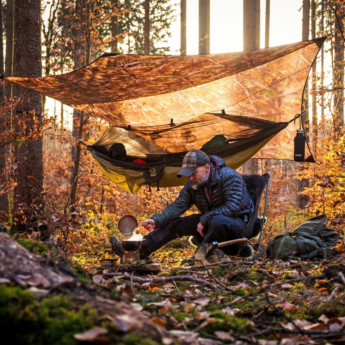 A man camping in a forest with a hammock suspended under a large camouflage tarp, cooking over an open fire while wearing a blue puffer jacket and plaid cap.