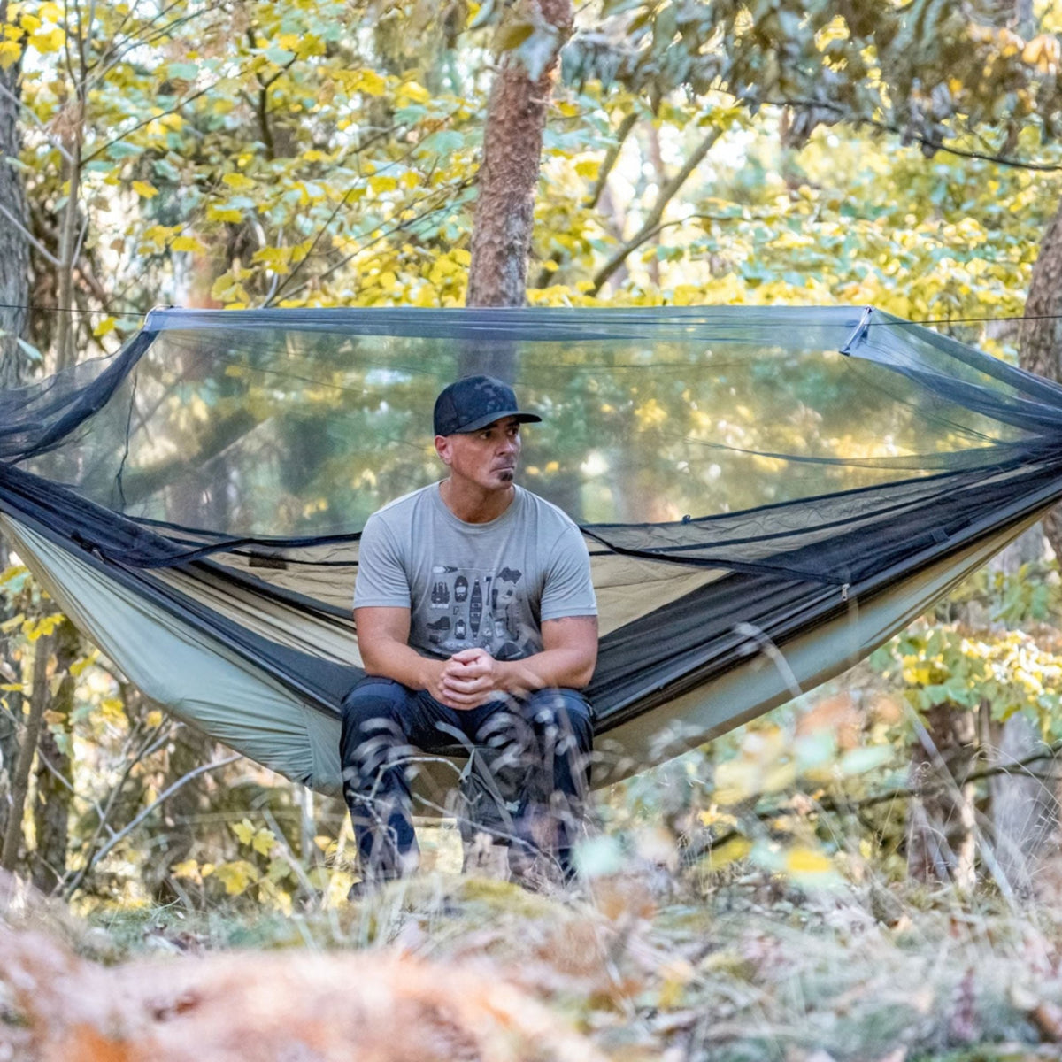 A man sits inside a large black mosquito net hammock suspended in a wooded forest setting.