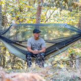 A man sits inside a large black mosquito net hammock suspended in a wooded forest setting.
