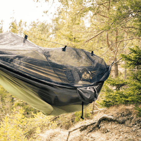 A man lies inside a black mosquito net hammock suspended between trees in a forest setting.